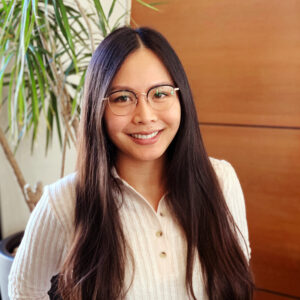 Karen Leung in a cream colored sweater, with long dark hair, standing in front of a wooden panel and plant.