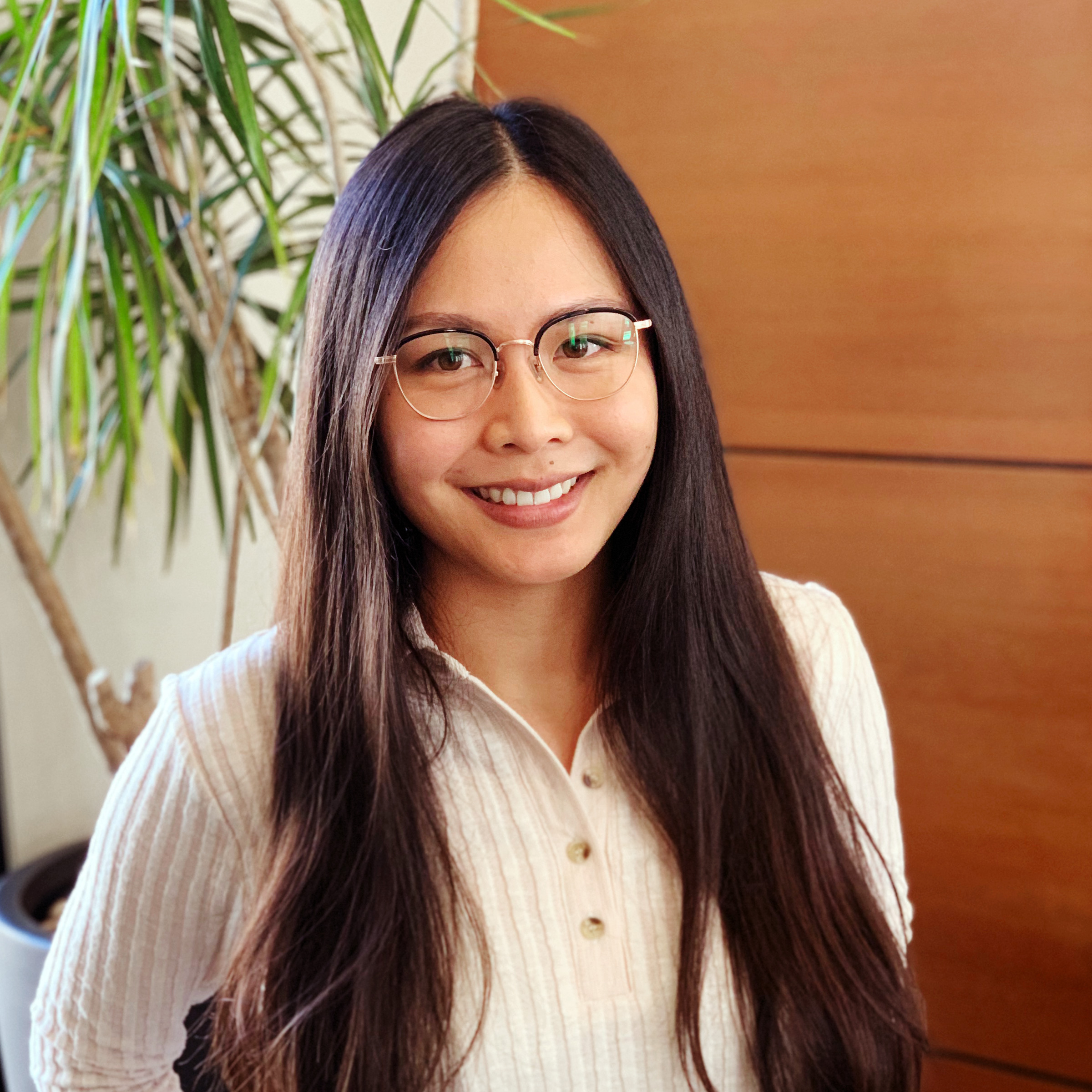 Karen Leung in a cream colored sweater, with long dark hair, standing in front of a wooden panel and plant. 